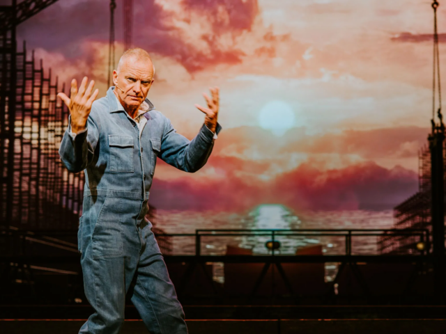 Sting as Jackie White, in a denim boiler suit, raises both hands mid-song against a projected sunset over the Tyne.