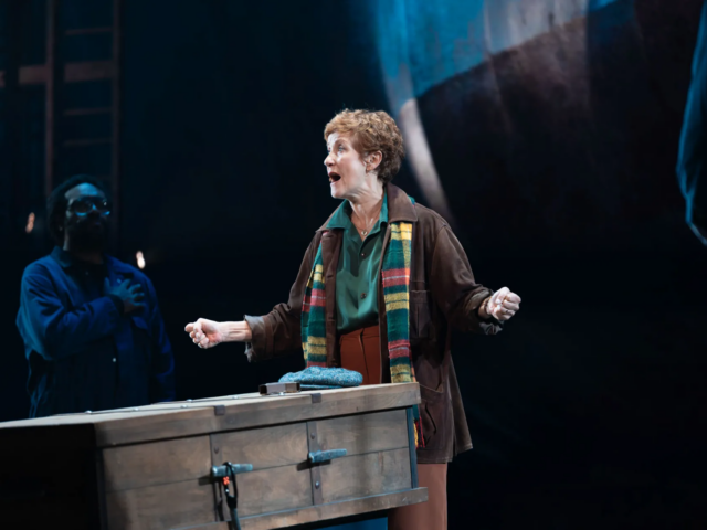 Peggy White sings beside a workbench, holding a hammer and a flat cap.