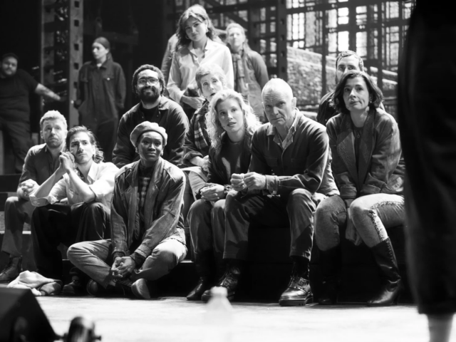 Black and white: the company seated together on the stage steps during rehearsals, shipyard scaffolding behind them.