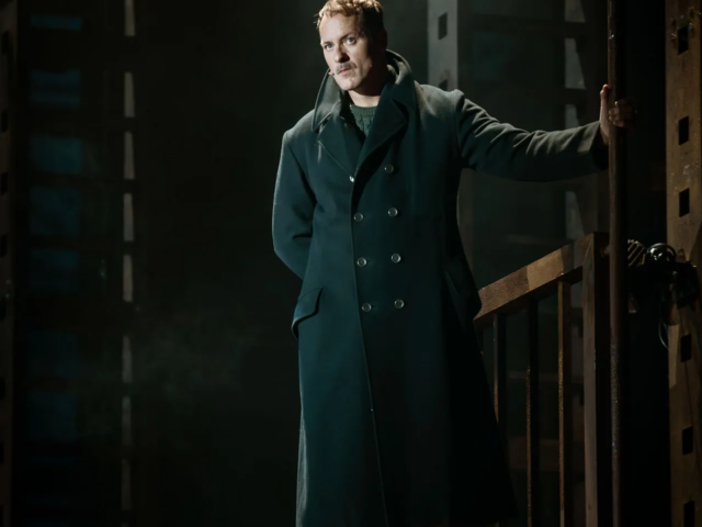 A man in a long dark overcoat stands on a raised platform, one hand on a metal rail, lit against the shipyard steelwork.