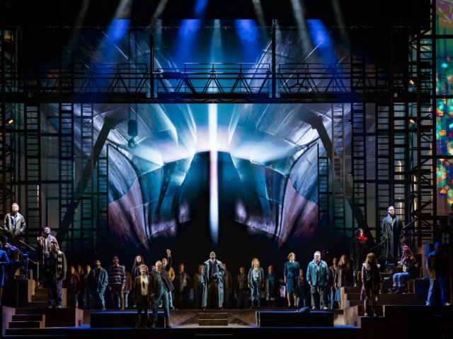 The full company across the stage beneath a vast projection of a ship's bow lit from behind.