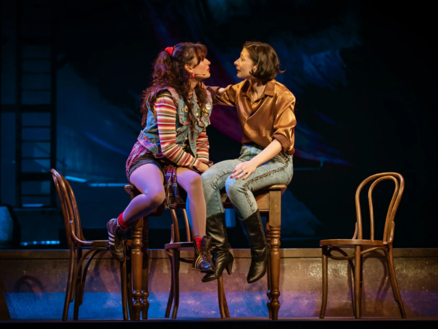 Ellen and Meg sit facing each other on wooden chairs, mid-scene.