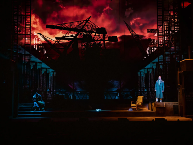 The full stage lit in red, silhouetted shipyard cranes overhead, a lone figure in a long coat on a raised platform.