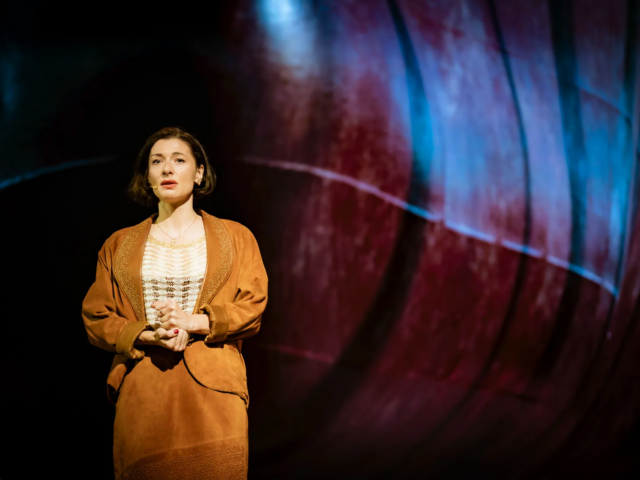 Meg stands alone in a tan suede jacket and skirt, hands clasped, against a projected ship's hull.