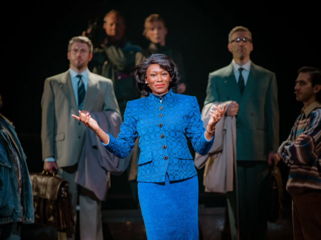 Baroness Tynedale, in a blue check suit, addresses the yard with her hands open, suited managers behind her.