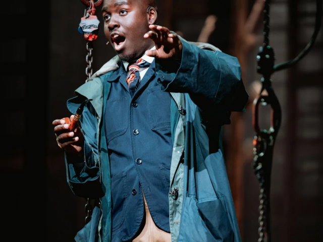 The Wallsend Ferryman, in a teal raincoat and tie, reaches out mid-song beside a hanging crane hook.