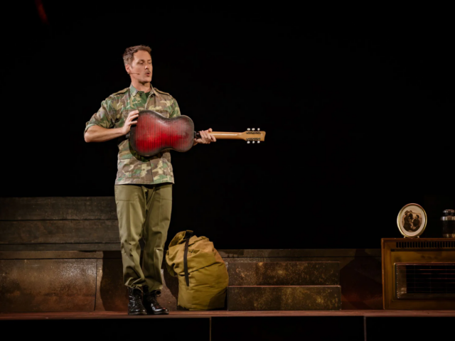A man in a camouflage jacket holds a battered red guitar out in front of him, a kitbag at his feet.