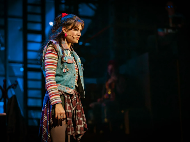 Ellen stands in profile in a badge-covered denim waistcoat and striped top, lit blue against the shipyard.