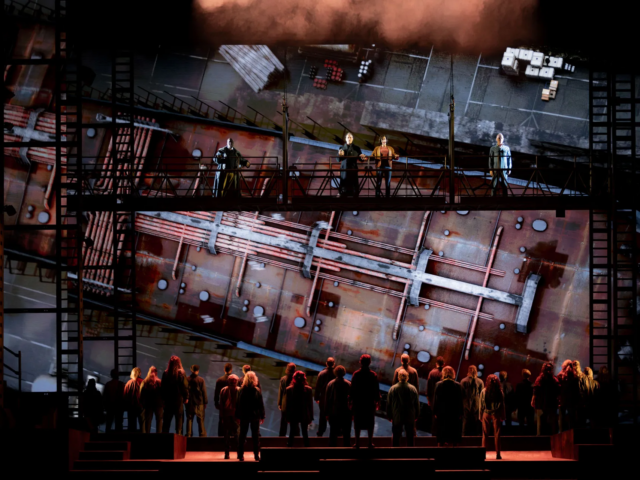 The company face upstage towards a vast projection of a rusting hull, four figures watching from the gantry above.