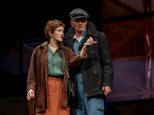 Jackie White, in a flat cap and pea coat, stands behind Peggy as she points into the distance.