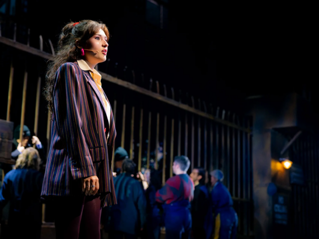 Ellen in an oversized pinstriped blazer, lit apart from the crowd behind the shipyard railings.