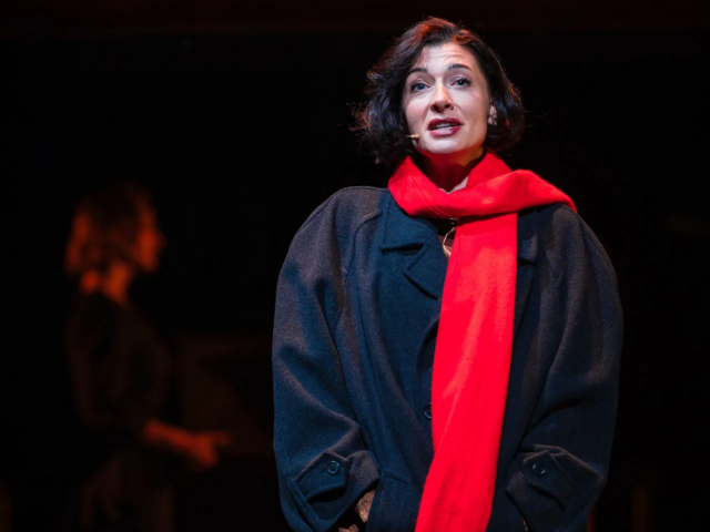 Meg in a dark overcoat and red scarf, alone in a spotlight.