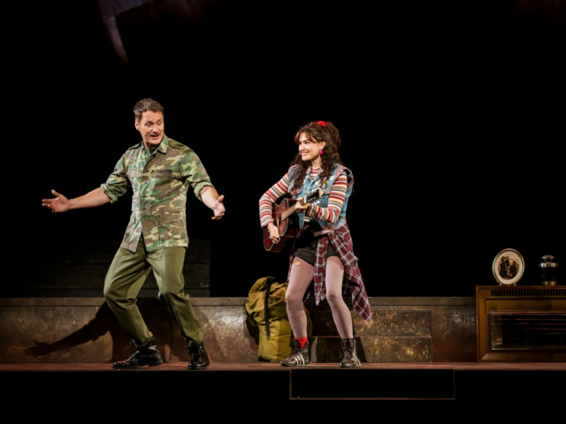 A man in a camouflage jacket dances as Ellen plays guitar beside him.