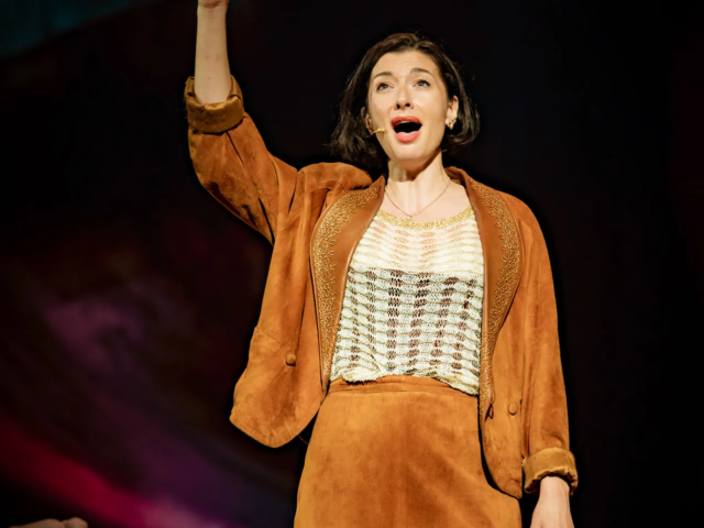 Meg sings with one arm raised, in a tan suede jacket and skirt.